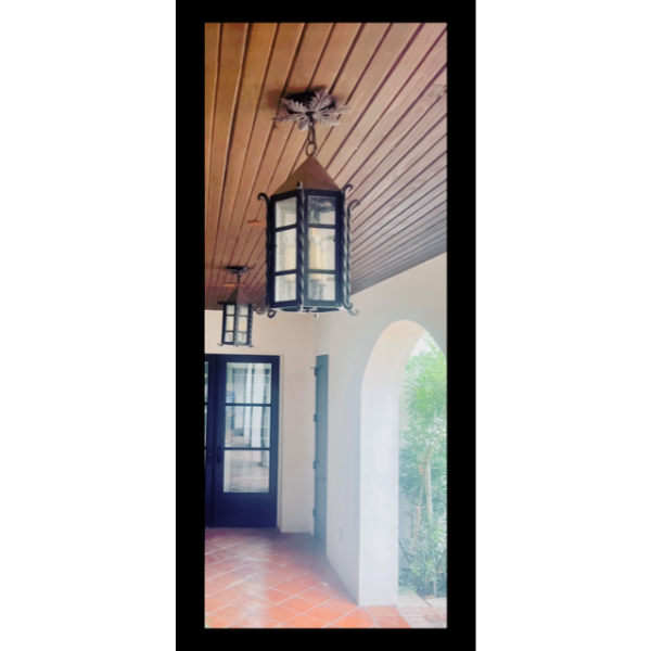 A striking wrought iron pendant light hangs from a wooden ceiling, featuring a geometric glass design. The fixture's elegant curves complement the surrounding clean white walls and terracotta tile floor, enhancing the warm, inviting atmosphere of the entryway.