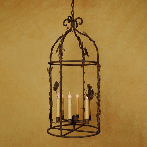 A decorative wrought iron chandelier featuring an elegant cage design adorned with intricate leaf motifs. It holds four candle-like bulbs, providing a warm, inviting glow. The fixture hangs from a chain against a textured golden wall, enhancing its rustic charm.