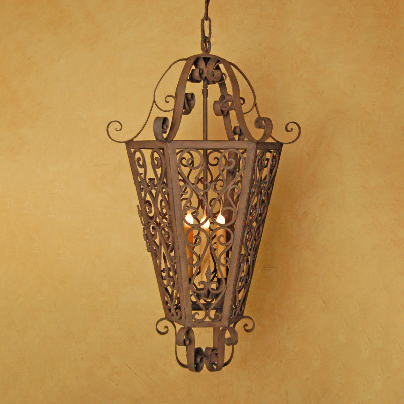 A beautifully crafted wrought iron pendant featuring intricate scrollwork and a warm, inviting glow from three inner lights, hanging against a textured ochre wall.