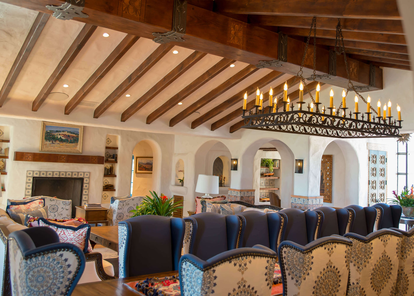 A close-up of a Spanish-style wrought iron chandelier with intricate scrollwork and candle-like lights, hanging from a wooden ceiling beam, highlighting its elegant design and rustic charm.