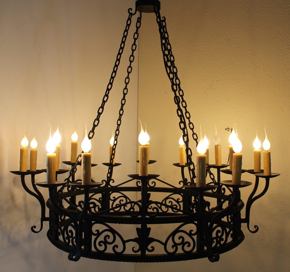 A large wrought iron chandelier featuring intricate scrollwork and candle-like lights. It hangs from a sturdy chain and provides an elegant, warm glow.