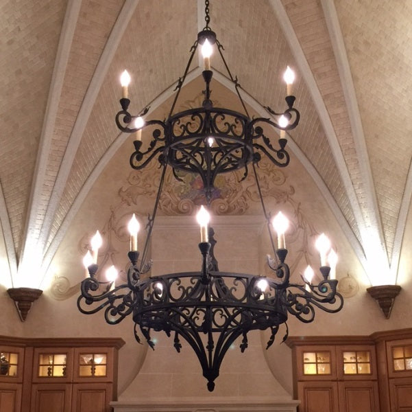 A large, intricately designed wrought iron chandelier hangs from a vaulted ceiling, showcasing ornate curves and elegantly positioned lights. The warm glow illuminates the sophisticated space, complementing the rich wooden cabinetry below.