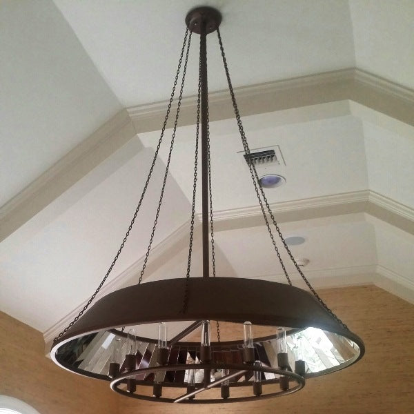 A large, circular wrought iron chandelier with a sleek, dark finish. It features multiple light bulbs arranged symmetrically and is suspended by four chains from the ceiling, adding an elegant touch to the room's high ceiling and architectural details.