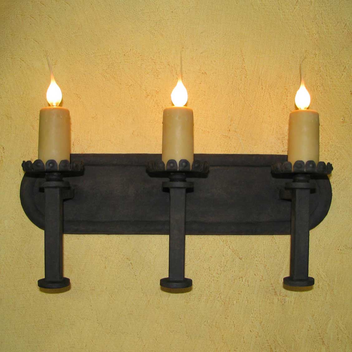 A sleek wrought iron wall sconce featuring three candle holders with warm, flickering LED lights, mounted against a textured yellow wall. The elegant design showcases ornate details and sturdy craftsmanship, enhancing any interior with a touch of sophistication.