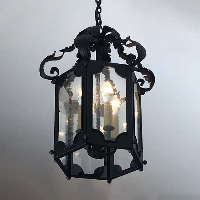 A beautifully crafted wrought iron pendant light featuring intricate scrollwork and a hexagonal shape. The fixture has clear glass panels showcasing multiple glowing bulbs inside, providing warm illumination.