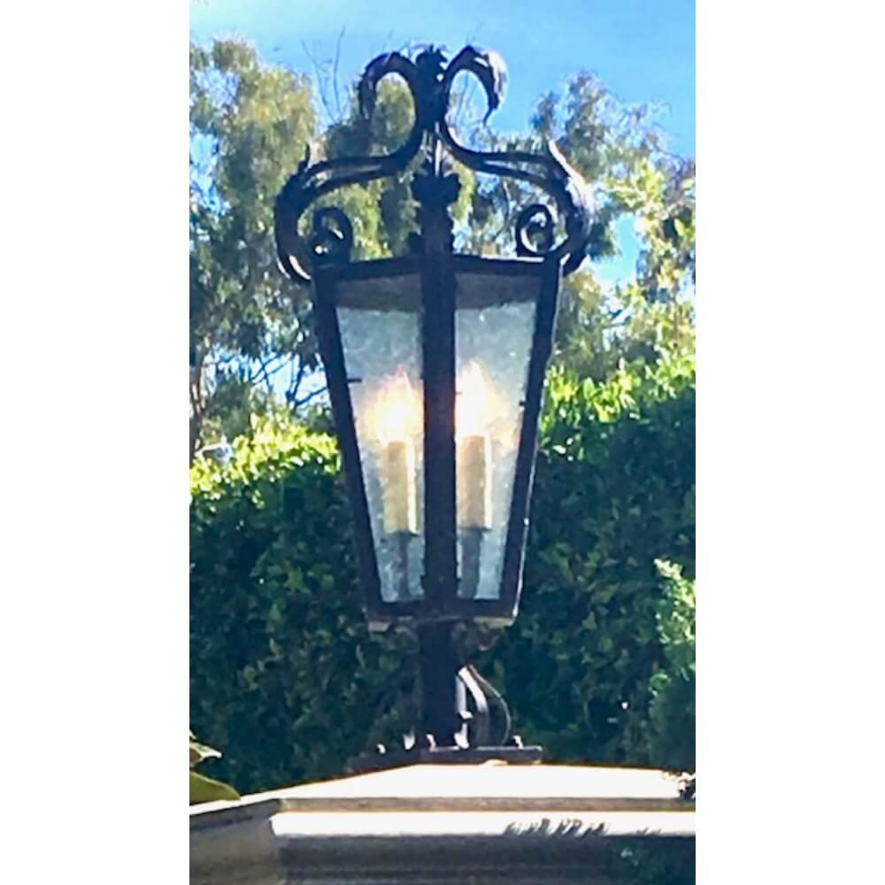 A decorative wrought iron sconce featuring a classic design with a distinct curved top. It showcases two candle-like bulbs behind textured glass, surrounded by a dark metallic frame. The backdrop includes lush greenery, enhancing the elegant appeal of the fixture.