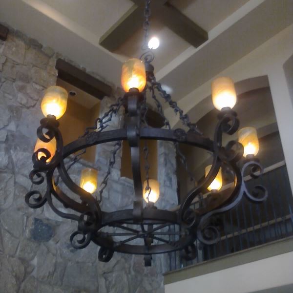 An ornate wrought iron chandelier featuring a circular design with curling accents. Eight golden-hued glass shades emit warm light, hanging from a sturdy chain. The backdrop showcases a rustic stone wall, enhancing the fixture's elegant charm.