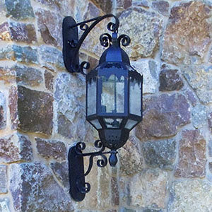 A decorative wall sconce featuring ornate wrought ironwork with intricate scroll designs, complementing a glass lantern. The fixture is mounted against a textured stone wall, showcasing its elegance and craftsmanship.