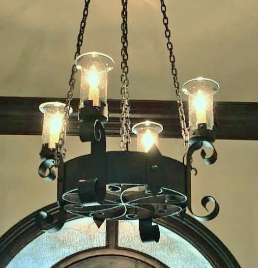 A beautifully crafted chandelier featuring a circular wrought iron design with decorative swirls. It holds multiple glass candle-style lights, suspended by chains from the ceiling, casting a warm glow in the room.