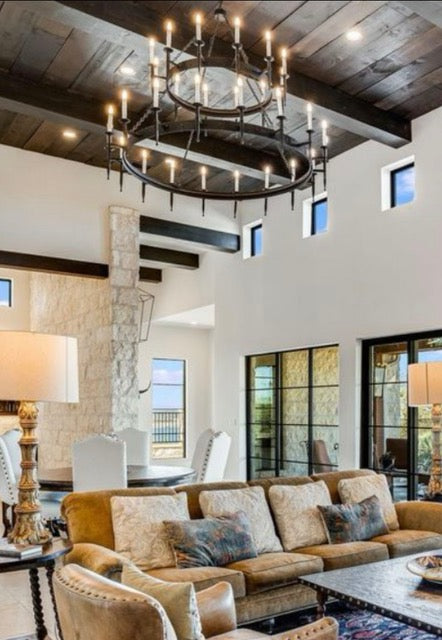 A large, rustic chandelier featuring multiple candle-like lights arranged in two circular tiers. The fixture hangs from a ceiling of wooden beams, adding warmth and elegance to the space.