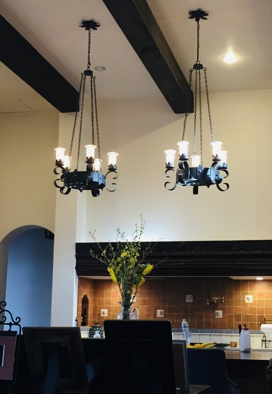 Two elegant wrought iron chandeliers hang from a ceiling with wooden beams. Each fixture features six candle-like lights, casting a warm glow over a kitchen area adorned with a simple floral arrangement and rustic decor.