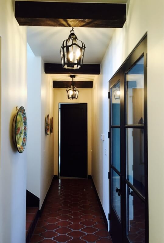 Three elegantly designed wrought iron fixtures hang from the ceiling in a warmly lit hallway. The space features terracotta tiles and decorative plates on the walls, leading to a black door and glass-paneled door. The fixtures add a touch of rustic charm and illumination to the interior design, enhancing the inviting ambiance of the corridor.