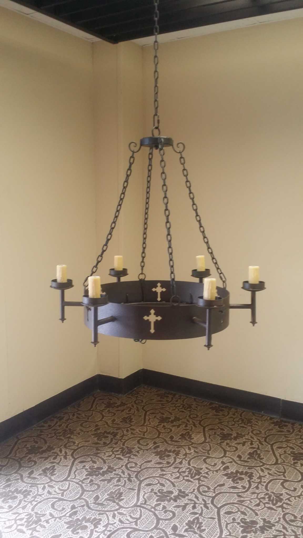 A handcrafted chandelier featuring a dark wrought iron finish and six candle-like light fixtures. It is adorned with decorative crosses, suspended from a sturdy chain, adding an elegant touch to any space. The background highlights a patterned floor.