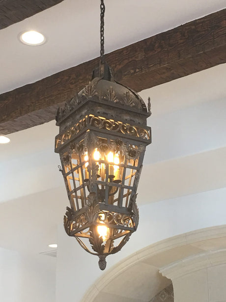 A beautifully crafted wrought iron pendant light featuring intricate floral designs and a rounded top. The fixture, hanging from a chain, illuminates the space with warm bulbs, showcasing its ornate details against a backdrop of wooden beams and soft white walls.