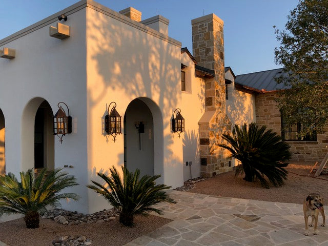 A beautifully crafted wrought iron sconce featuring a diamond-shaped top and glass panels. The warm glow from the interior lights illuminates the surrounding area, creating an inviting ambiance. Adjacent to a green shrub and mounted on a neutral wall, the design showcases elegant curves and sturdy craftsmanship, making it an ideal addition to any exterior space.