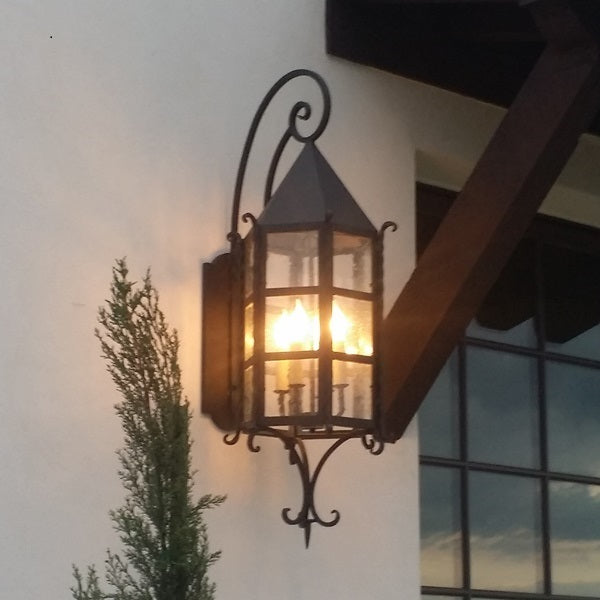 A beautifully crafted wrought iron sconce featuring a diamond-shaped top and glass panels. The warm glow from the interior lights illuminates the surrounding area, creating an inviting ambiance. Adjacent to a green shrub and mounted on a neutral wall, the design showcases elegant curves and sturdy craftsmanship, making it an ideal addition to any exterior space.