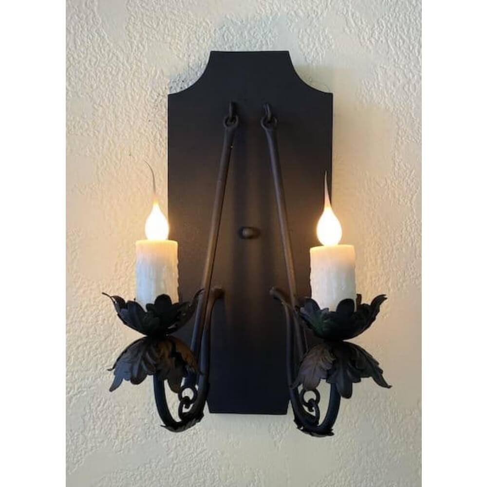 A decorative wall sconce featuring two candle-like bulbs surrounded by intricate wrought iron foliage. The fixture is mounted on a textured black backplate, adding elegance to any space.