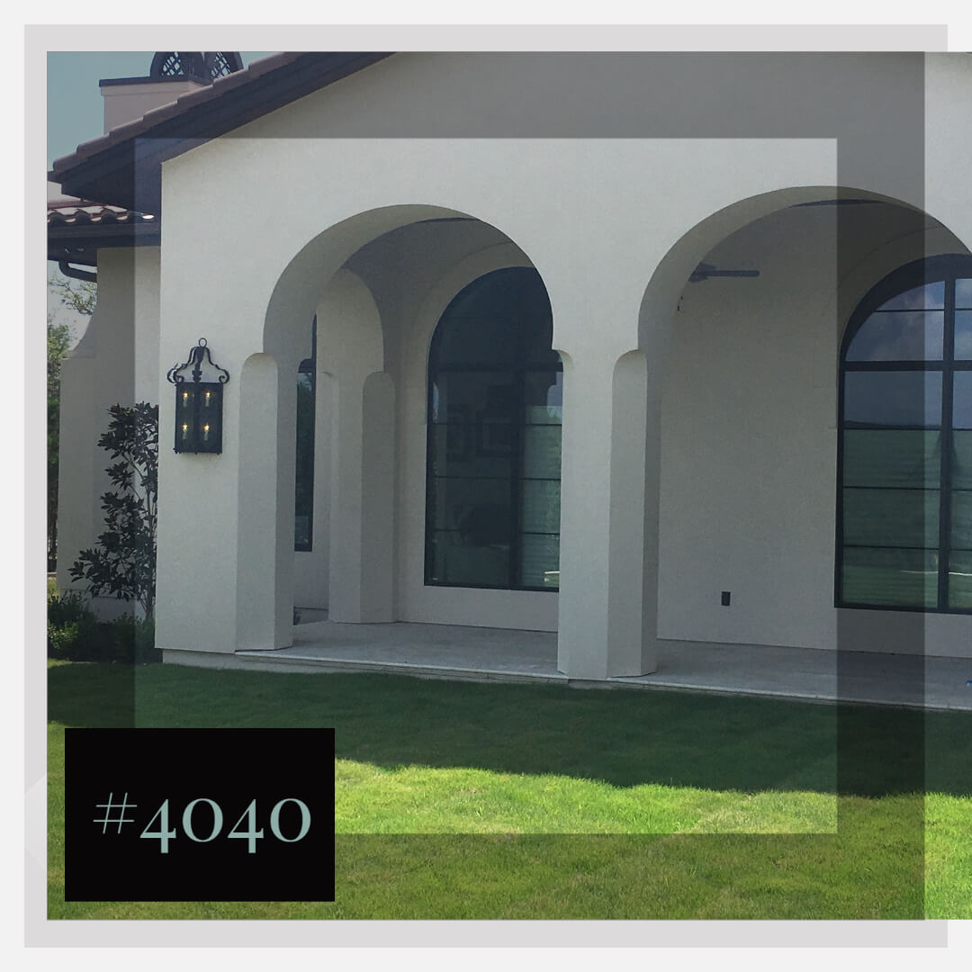 A decorative wrought iron sconce hangs on a white stucco wall beneath elegant arches. The fixture features multiple glowing lights, enhancing the outdoor space's charm and ambiance. Lush green grass surrounds the area, complementing the striking architectural details.