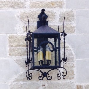 A decorative wall-mounted sconce featuring a black wrought iron frame with intricate scrollwork, housing three candle-like bulbs. The design includes a peaked roof and floral accents, complementing rustic or traditional settings.