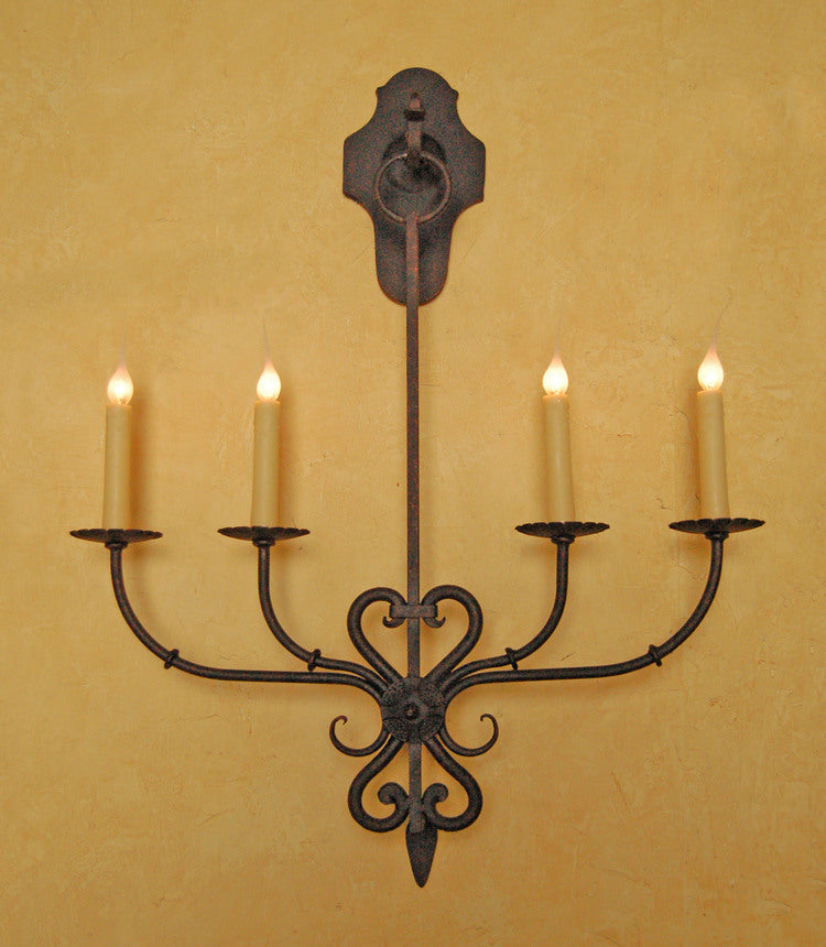 A decorative wrought iron wall sconce featuring intricate curves and elegant candle holders. The design highlights a classic style with four slender arms extending from a central point, topped with lighted candles, set against a warm, textured background.
