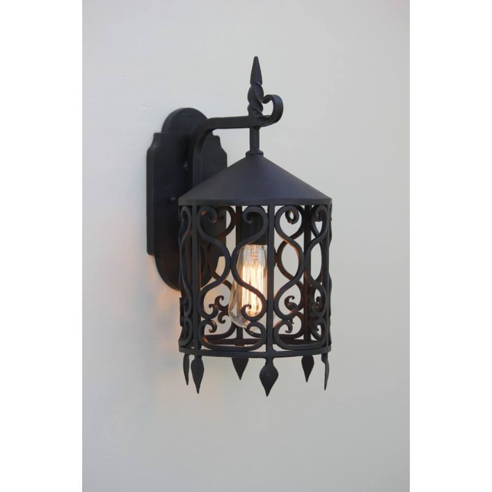 Wrought iron wall sconce featuring intricate scrollwork and a pointed top. The fixture holds a glowing bulb, enhancing ambient light and adding elegance to indoor or outdoor spaces.