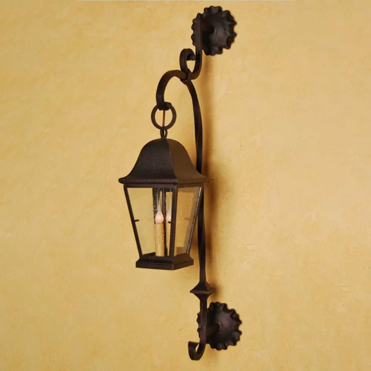A stylish wrought iron sconce featuring a decorative curved arm and a glass lantern with a classic conical top. The fixture showcases elegant craftsmanship and is mounted against a textured, warm-toned wall, adding charm and character to any space.