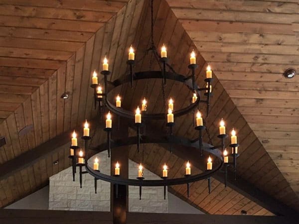 A large, rustic chandelier featuring multiple candle-like lights arranged in two circular tiers. The fixture hangs from a ceiling of wooden beams, adding warmth and elegance to the space.
