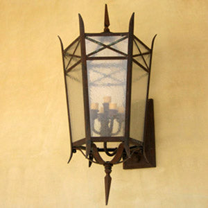 A decorative wrought iron sconce featuring an ornate design with a clear glass enclosure. The fixture showcases candle-like bulbs and intricate detailing, paired with a pointed top and elegant metalwork, ideal for enhancing any space's ambiance.