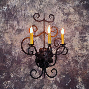 A beautifully crafted wrought iron sconce featuring intricate scrollwork and three candle-like lights, casting a warm glow against a textured backdrop.