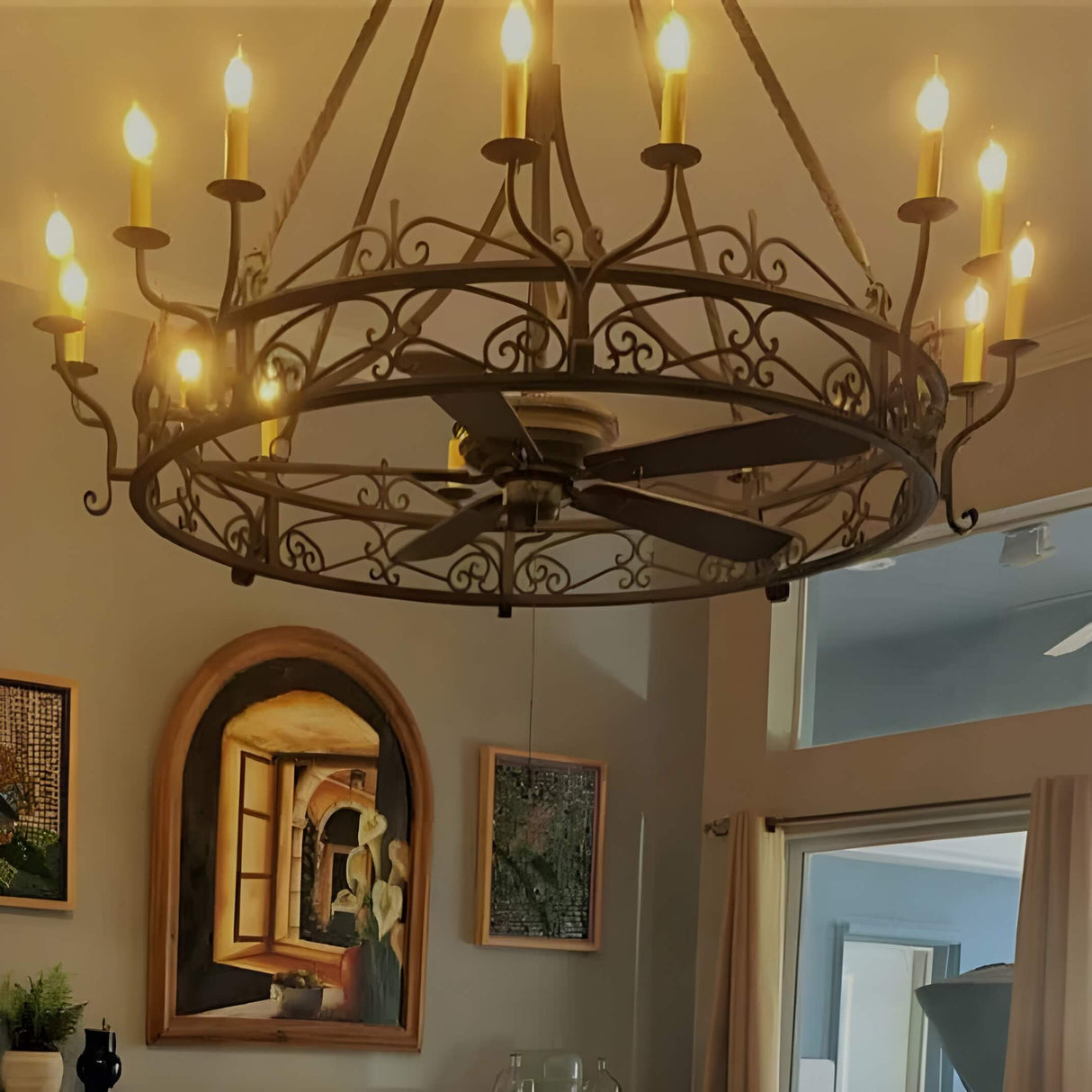 A large, ornate wrought iron chandelier with multiple candle-style lights hangs from the ceiling in a stylish room. The fixture features elegant swirls and curves, enhancing the warm ambiance of the space, which is adorned with framed artwork and decorative elements.