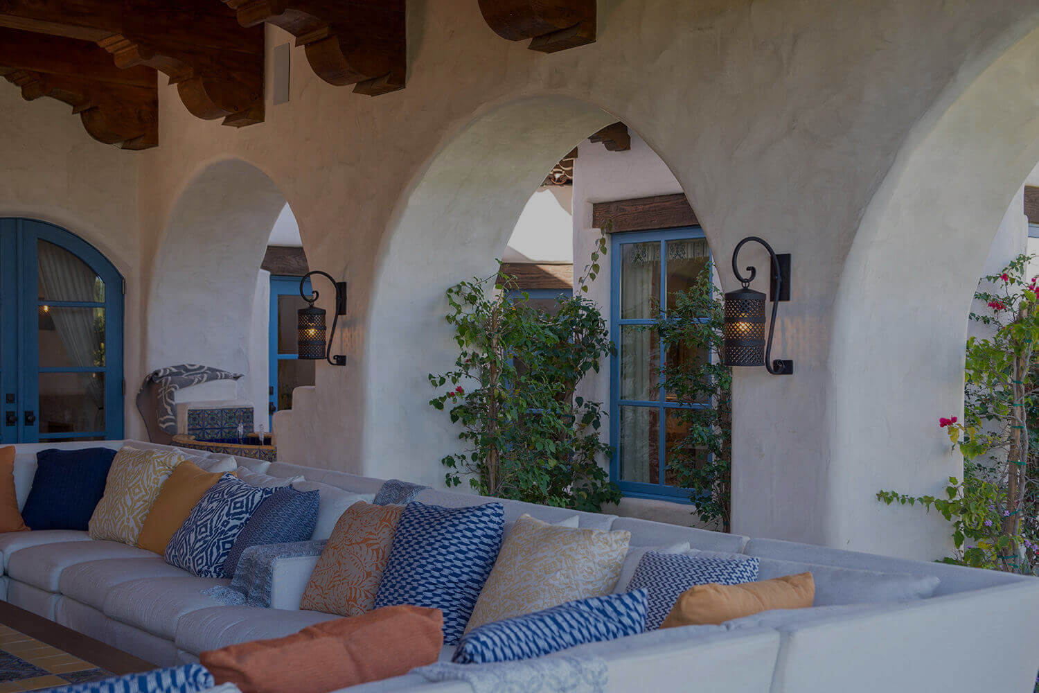 Wrought iron sconces illuminate a stylish outdoor space, complementing a cozy sofa adorned with colorful cushions. Arched walls and blue accents enhance the aesthetic.