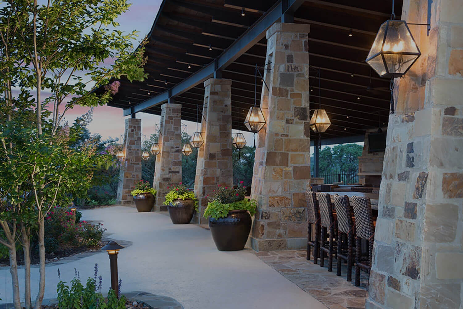 Illuminaries Custom Wrought Iron Lighting features elegant hanging fixtures that cast a warm glow, complemented by rustic stone pillars and lush greenery.