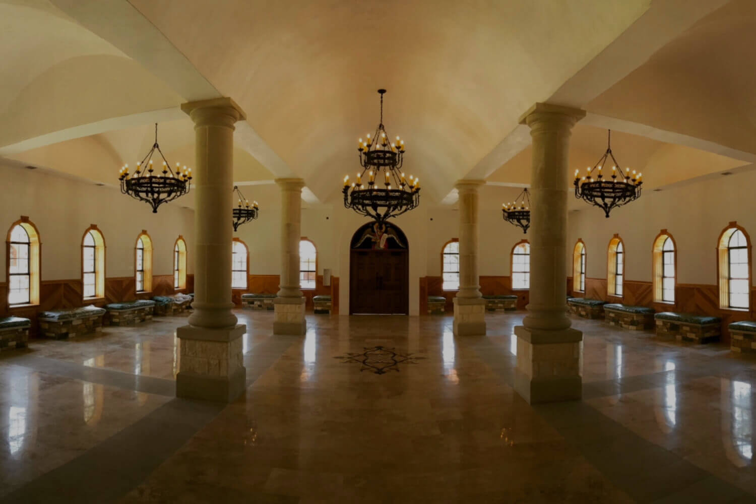 An elegant interior space with high ceilings features striking wrought iron chandeliers hanging from above. Columned architecture adds grandeur, while wooden benches line the walls, creating a warm, inviting atmosphere. The soft glow from the chandeliers illuminates the polished stone floor, enhancing the room's sophisticated design.
