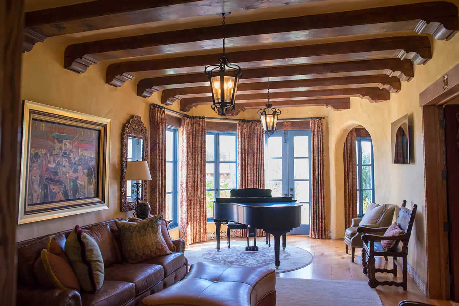 A cozy living room featuring elegant wrought iron pendant lighting, warm wooden beams, a grand piano, and plush seating. The windows are dressed with patterned curtains, complementing the soft tones of the walls. A beautifully framed artwork adds an artistic touch to the space.