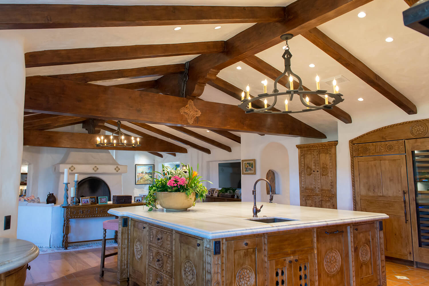 A stylish wrought iron chandelier hangs from wooden beams above a spacious kitchen island, enhancing the warm, rustic ambiance of the room.