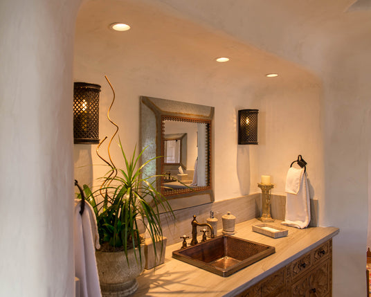 A beautifully lit bathroom featuring elegant wrought iron sconces on the walls, providing warm ambient light. The space is enhanced by a decorative mirror, lush greenery, and a stylish sink area.