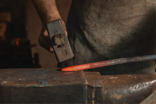 A close-up of a blacksmith's hand holding a hammer, poised to strike a glowing red iron rod resting on a heavy anvil. The workspace features a rustic environment, hinting at traditional craftsmanship. The anvil displays wear and tear, emphasizing the artistry behind custom wrought iron creations.