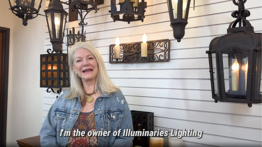 A woman stands in front of a display featuring various wrought iron lighting fixtures, including elegant chandeliers, decorative sconces, and unique pendants. The background showcases an assortment of designs, blending classic and contemporary elements. The woman smiles as she introduces herself as the owner of Illuminaries Lighting.