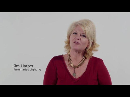 A woman with blonde hair speaks in front of a plain background. She represents Illuminaries Custom Wrought Iron Lighting, showcasing their unique wrought iron lighting fixtures that blend elegance and craftsmanship.