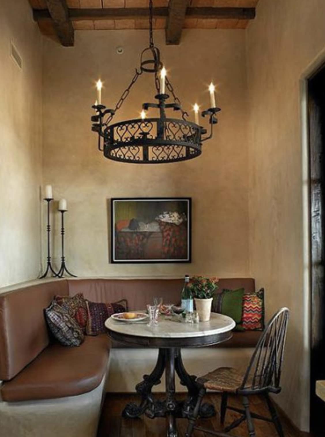 A beautifully designed wrought iron chandelier hangs above a round, marble dining table in a cozy nook. The fixture features candle-like bulbs and intricate scrolling patterns, complementing the earthy tones of the walls and wooden beams. A plush, cushioned seating area is adorned with colorful pillows, while a small vase filled with flowers and glasses of water add a charming touch to the setting. The ambiance exudes warmth and elegance, perfect for intimate dining.