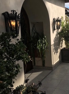 Two elegant wrought iron sconces flank an arched doorway, casting a warm glow. The fixtures feature intricate detailing and a classic design, enhancing the charm of the entryway adorned with lush greenery and soft lighting.