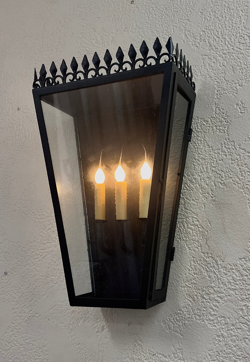 A wrought iron wall sconce featuring a pointed decorative top, clear glass panels, and three candle-style bulbs that emit a warm glow.