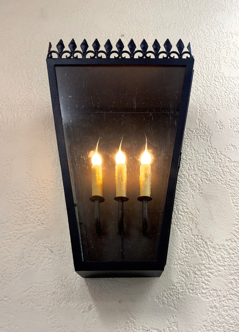 A wall sconce featuring three flickering candle-like lights enclosed in a sleek design with a decorative black iron top. The fixture casts a warm glow, creating an inviting atmosphere.
