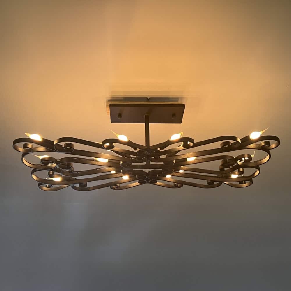 A decorative wrought iron chandelier featuring intricate, curling designs and multiple light sources. The fixture is mounted flush to the ceiling, casting a warm glow across the surrounding space.