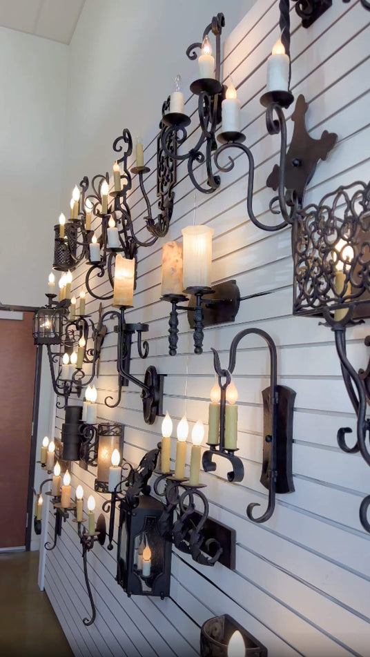 A wall display featuring a variety of custom wrought iron sconces, showcasing intricate designs, candle-style lights, and elegant finishes. The fixtures emit a warm glow, enhancing a charming atmosphere.