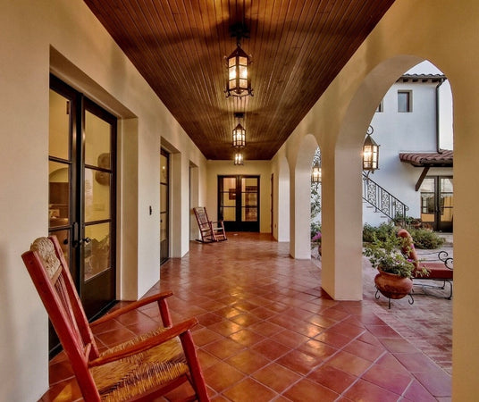 A cozy outdoor corridor featuring elegant wrought iron lighting fixtures hanging from a wooden ceiling. The rustic design complements the terracotta tiled floor and soft archways, creating an inviting atmosphere. Two wooden rocking chairs sit on one side, enhancing the warm, welcoming feel of the space.