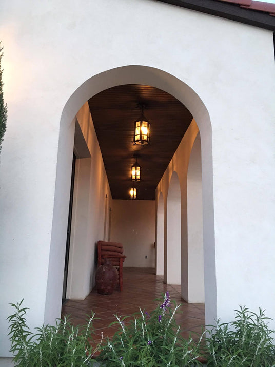 A view of a welcoming entryway showcasing three elegant wrought iron fixtures hanging from a wooden ceiling. The arches and white walls accentuate the ambiance, while a rustic pot and a bench with rolled towels add charm to the setting. Lush greenery with purple flowers frames the scene, enhancing the inviting atmosphere.