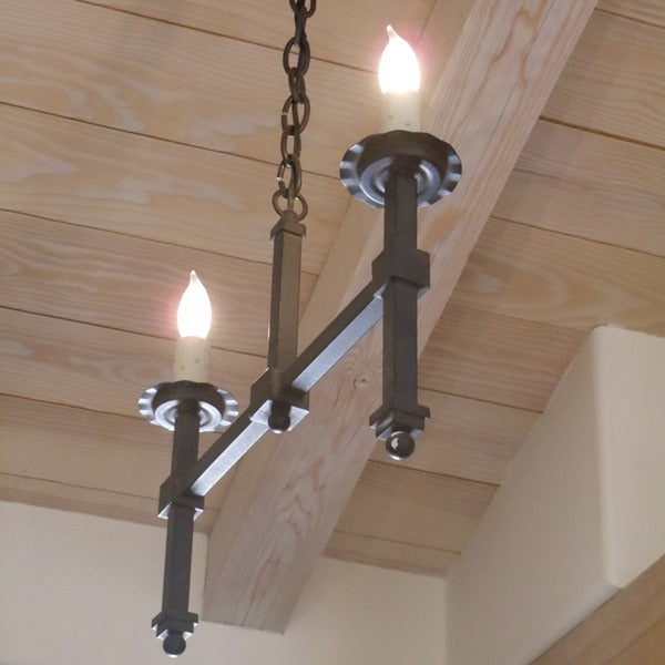A two-light wrought iron chandelier featuring elegant, candle-like bulbs and a sleek, linear design. The fixture hangs from a chain against a wooden ceiling, adding sophistication to any space.