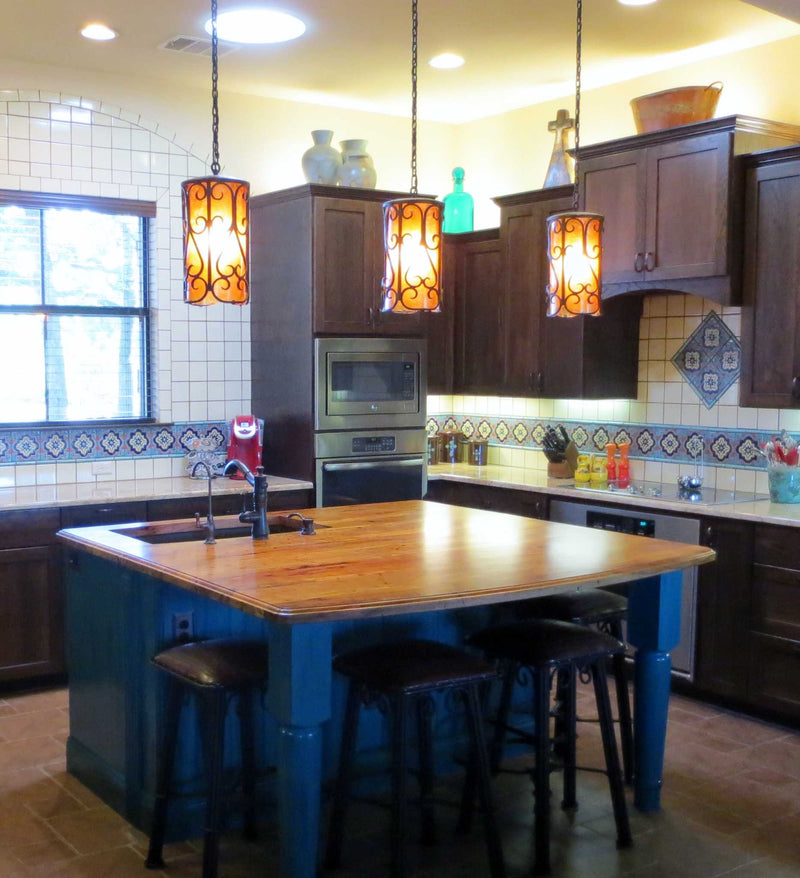 Three pendant lights hang above a wooden kitchen island, illuminating the space with warm, inviting light. The fixtures feature intricate wrought iron designs complemented by amber-colored glass shades, adding a touch of elegance to the rustic kitchen decor. Dark cabinetry and decorative tile work enhance the overall aesthetic.