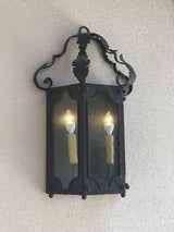 A beautifully crafted wrought iron sconce featuring intricate scrollwork and a decorative top. Dual glass panels enclose two glowing bulbs, enhancing its classic elegance.